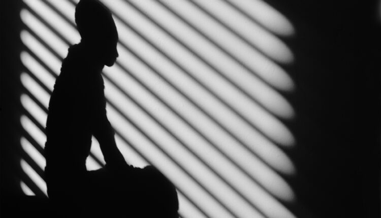 A photo of shadows on a wall shows the silhouette of a person sitting superimposed with the shadows of bars on a window behind them.
