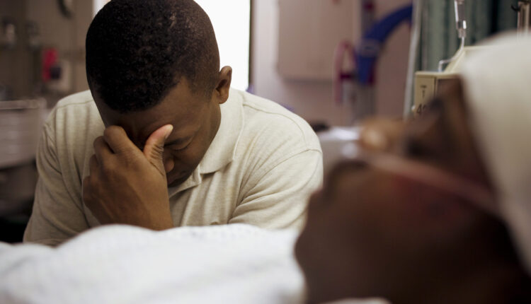 A photo of a Black man sitting beside his son in a hospital bed, covering his face.