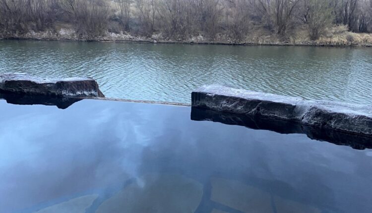 A hot springs pool overlooks the river, with shrubs on the shore and mountains in the distance under a cloudy sky.