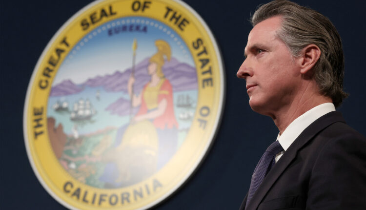 A photo of Governor Gavin Newsom during a press conference taken from the side. On the wall behind him is California's state seal.