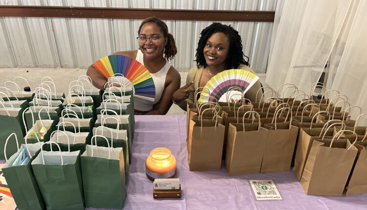 Two college students, Limya Harvey (left) and Cydney Mumford (right), sit at a table covered with small, paper grab-bags. The women smile towards the camera, each holding a paper fan with pride-flag colors.