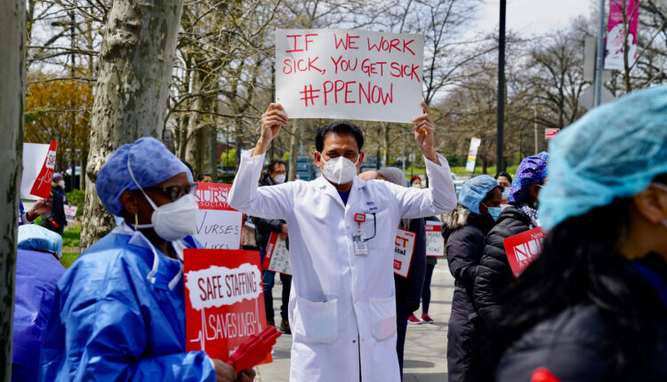 Public health workers, doctors, and nurses protest outside a New York City hospital, holding signs. A doctor, wearing a face mask and white coat, is photographed in the center of a crowd. He holds a sign that reads, "If we works ick, you get sick. #PPENOW"