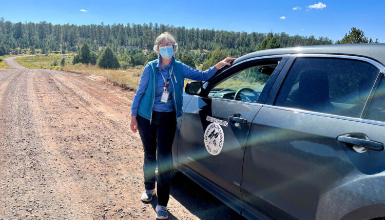 A woman in a blue shirt and mask stands in front of a car parked along a dirt road