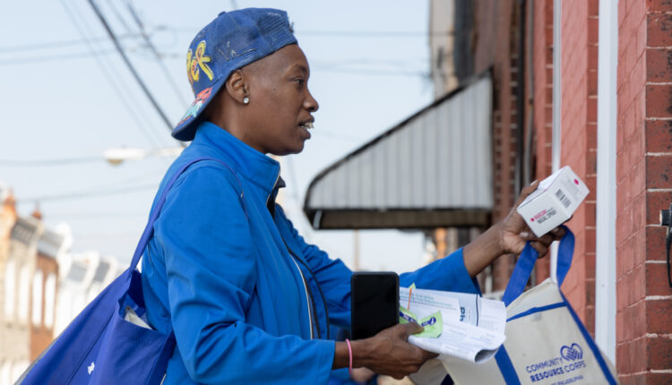 A woman in a backwards blue cap and matching blue sweat jacket hands out Narcan door to door