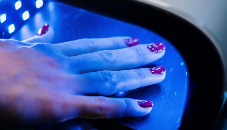 A person with painted nails holds their hand under machine emitting a blue light.
