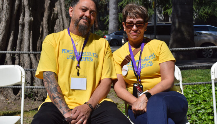 A photo of a man and a woman sitting next to each other outside. They both are wearing yellow shirts that read, "People with disabilities deserve services."