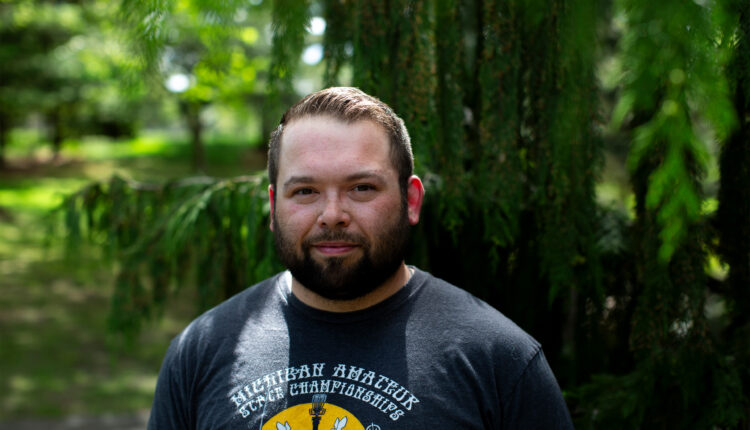 A man with brown hair and bear stands outside by a tree and looks at the camera.