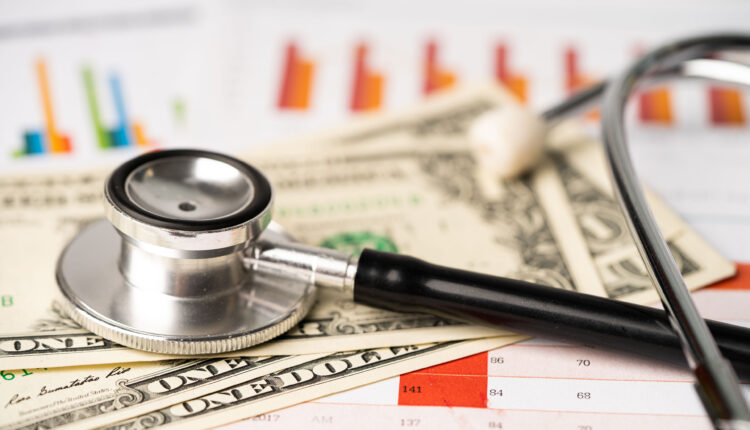 A stethoscope rests on top of a pile of U.S. dollar bills and finance report papers.